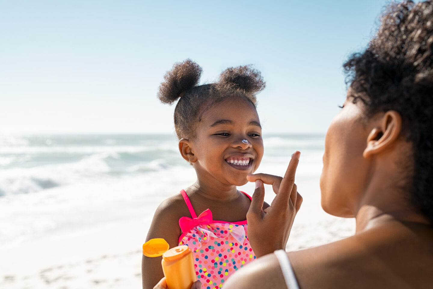 Black mother applying sunscreen on smiling little Black girl with the ocean and beach behind them.