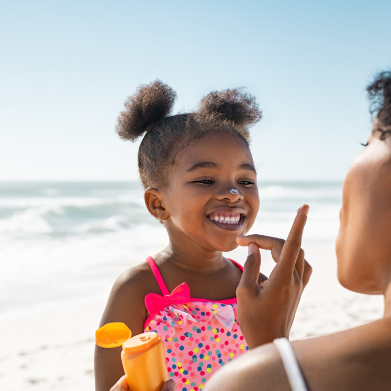 Black mother applying sunscreen on smiling little Black girl with the ocean and beach behind them.