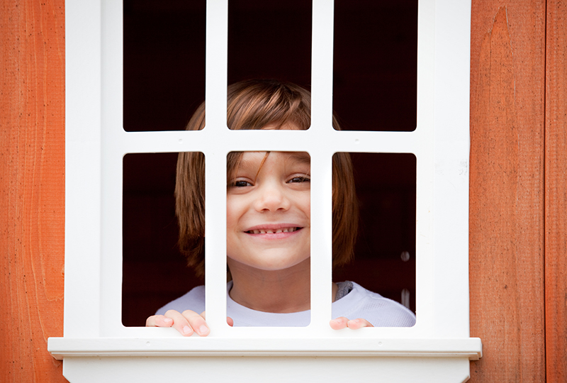 A young boy smiles as he peeks though the window of his playhouse.
