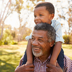 Older African American man carries a young boy on his shoulders outdoors on a sunny day.