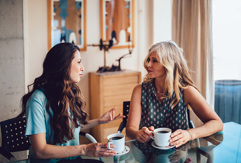 Two women talk while sitting together at a kitchen table drinking coffee