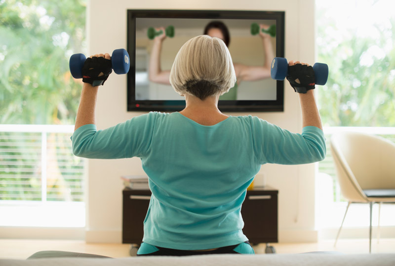 View of older woman from back as she lifts dumbbells while watching an exercise video