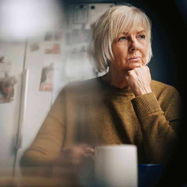 An older white woman sits at a table with a coffee mug looking stressed.