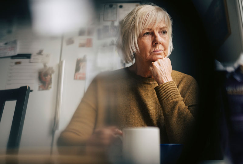 An older white woman sits at a table with a coffee mug looking stressed.