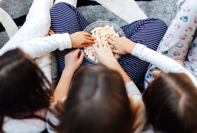 Overhead shot of three grade-school aged girls wearing pajamas sharing a bowl of popcorn at a sleepver.