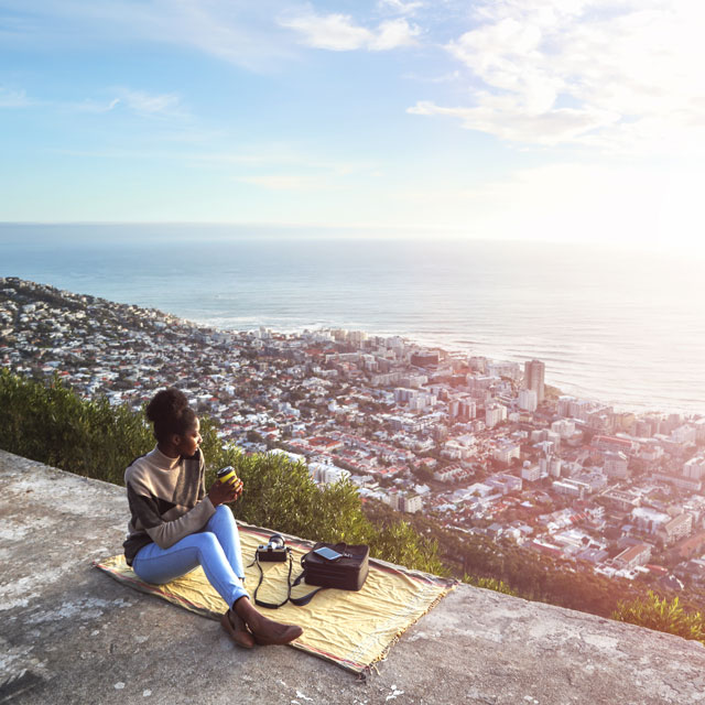 A woman sits on an overlook with the view below of a large city she has traveled to.