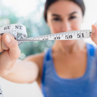 Woman holds up tape measure in foreground. Thumbnail sized image.