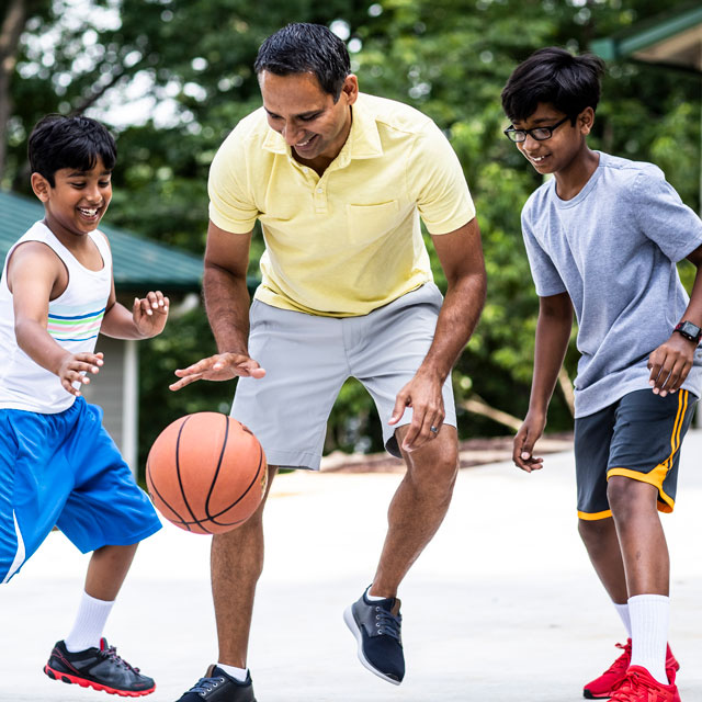 A father and two sons of Indian-American descent play basketball in a driveway.