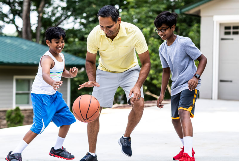 A father and two sons of Indian-American descent play basketball in a driveway.