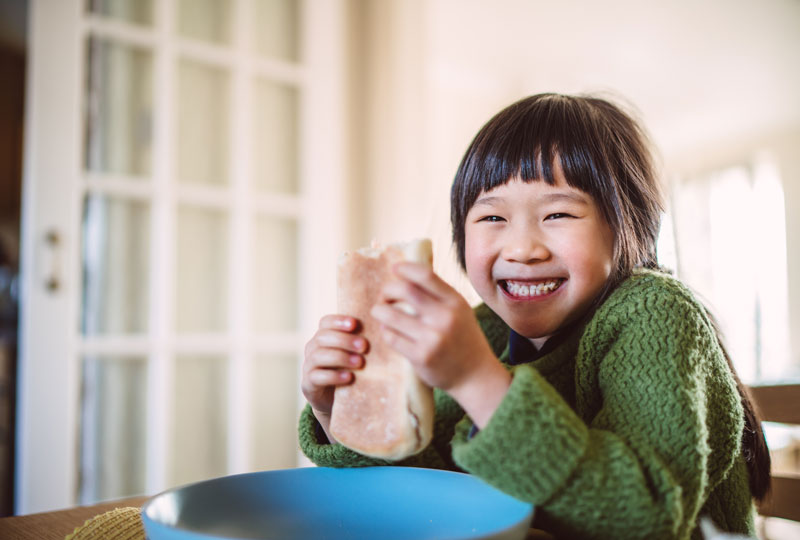A school-aged Asian girl eats a breakfast burrito sitting at a table at home.