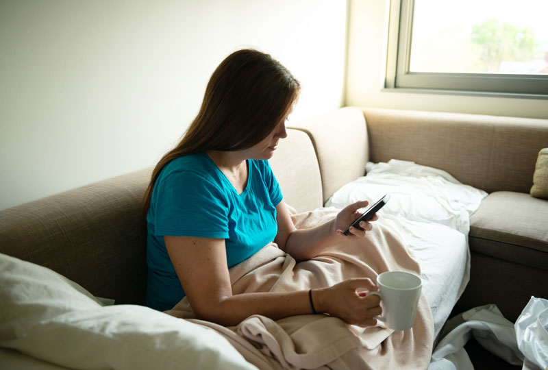 Woman sits on couch with blankets, holding cell phone and a mug of coffee.