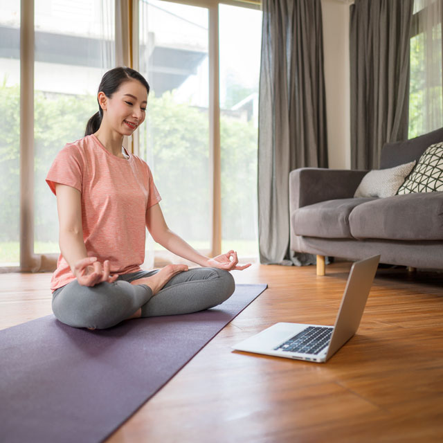 Young Asian woman sits in lotus position on a yoga mat at home with a laptop.