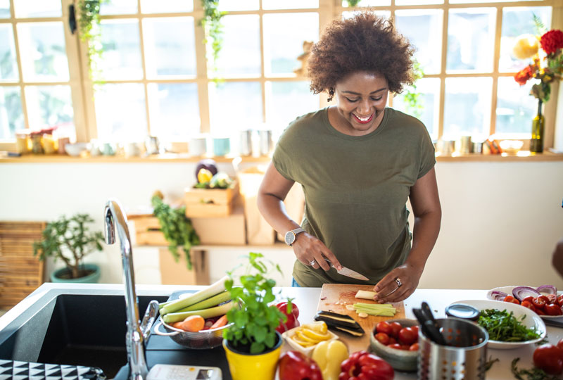 An African-American woman chops fresh vegetables in a sunny kitchen.