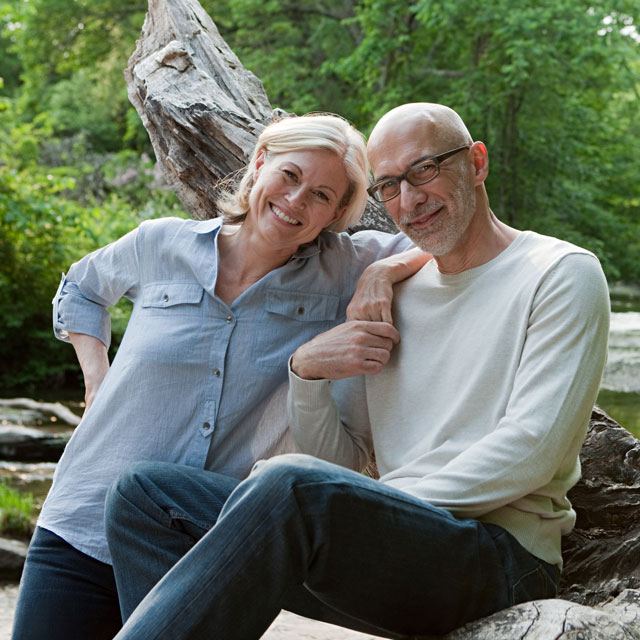 An older white man and woman sit on a log by the side of a river.