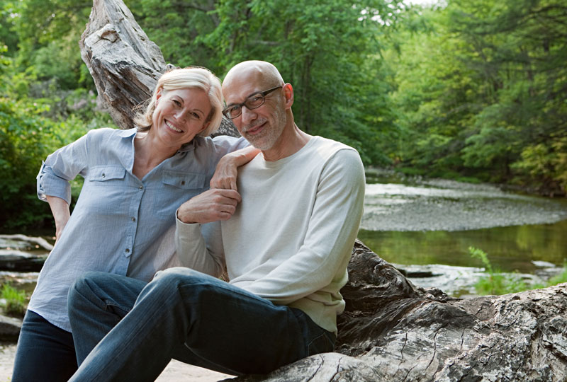 An older white man and woman sit on a log by the side of a river.