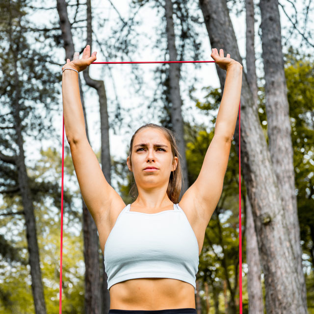 A young woman does a shoulder press with a resistance band in an outdoor workout.