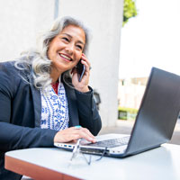 Older Latina woman works at a laptop and cell phone.