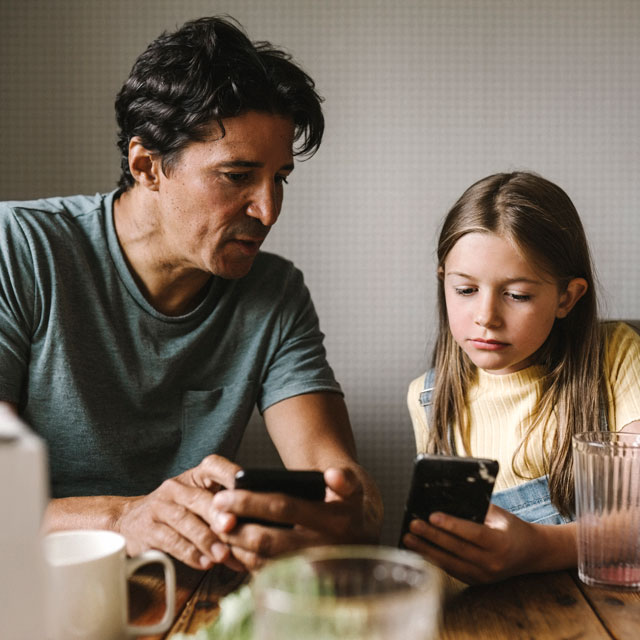 A father sits at the kitchen table with his school-aged daughter, talking and both holding cell phones.