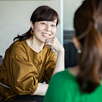 A view of a woman facing the camera, sitting, smiling and talking to a woman whose back is to the camera.