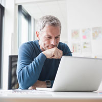 Middle-aged man sitting at a desk looking at his laptop screen in a sunny room.