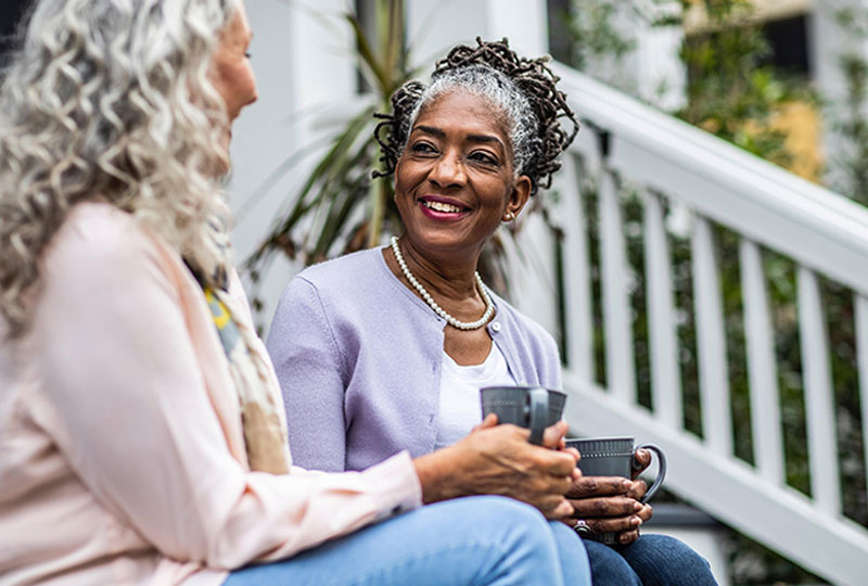 Two middle aged African American women sit on a stoop talking and holding coffee mugs.