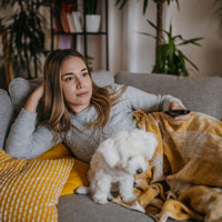 A young woman lies on a couch under a blanket, holding a TV remote, with a Maltese puppy by her side.