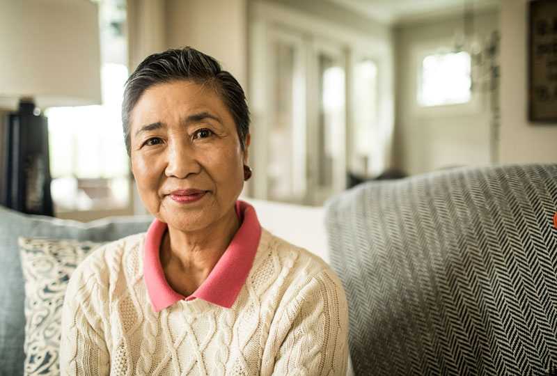 A head and shoulders shot of an older Asian woman sitting on a couch with a big cozy pillow.