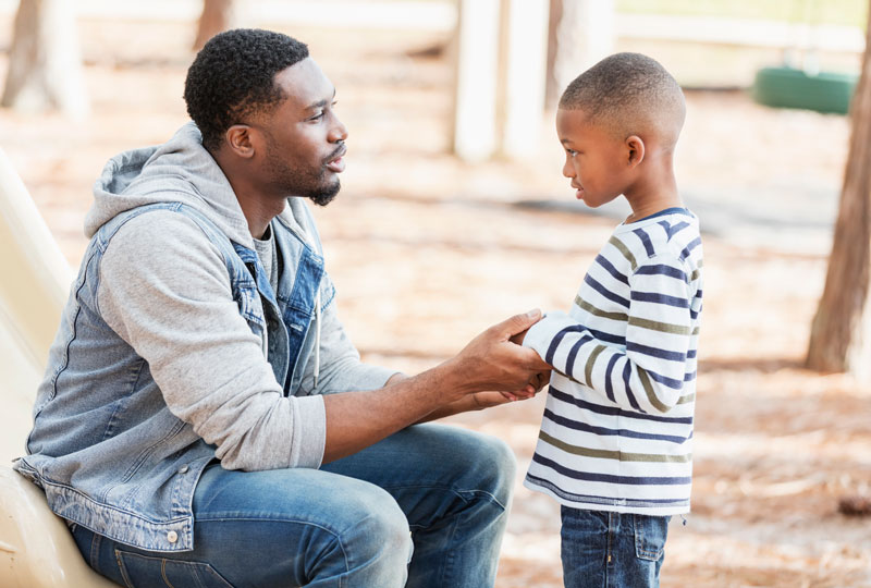 A young Black father sits outdoors talking with a school-aged boy.