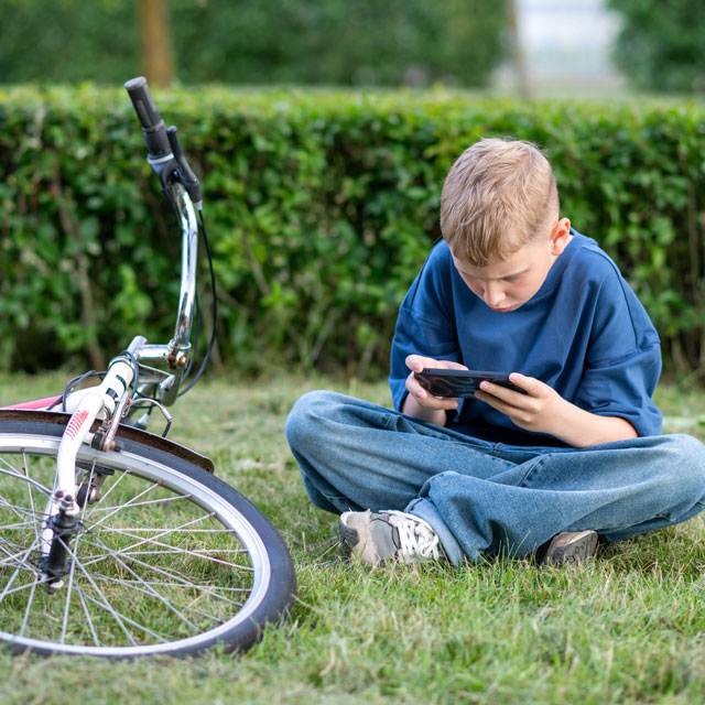 A tween boy sits outdoors on grass next to a bicycle, playing with a mobile phone.