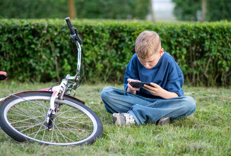 A tween boy sits on grass next to a bicycle, playing with a mobile phone.