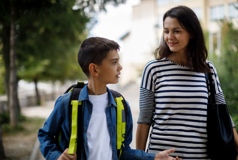 A young mother and her school-aged son walk along a shady sidewalk having a conversation.