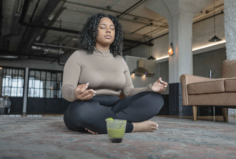 A young Black woman sits on a carpet in a large room, in a cross-legged yoga pose.