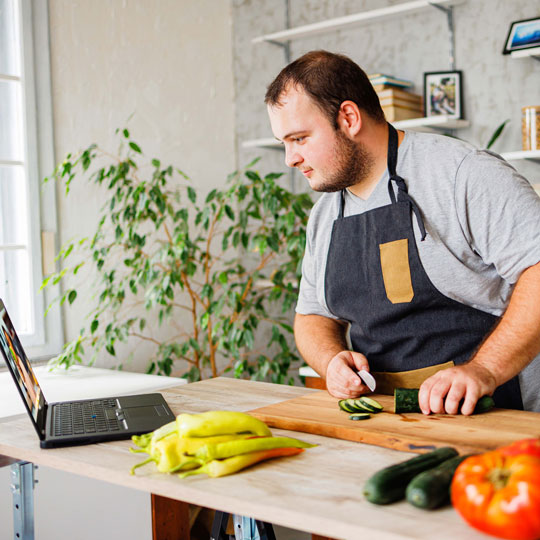 A man works in a kitchen preparing vegetables while following instructions on a laptop.