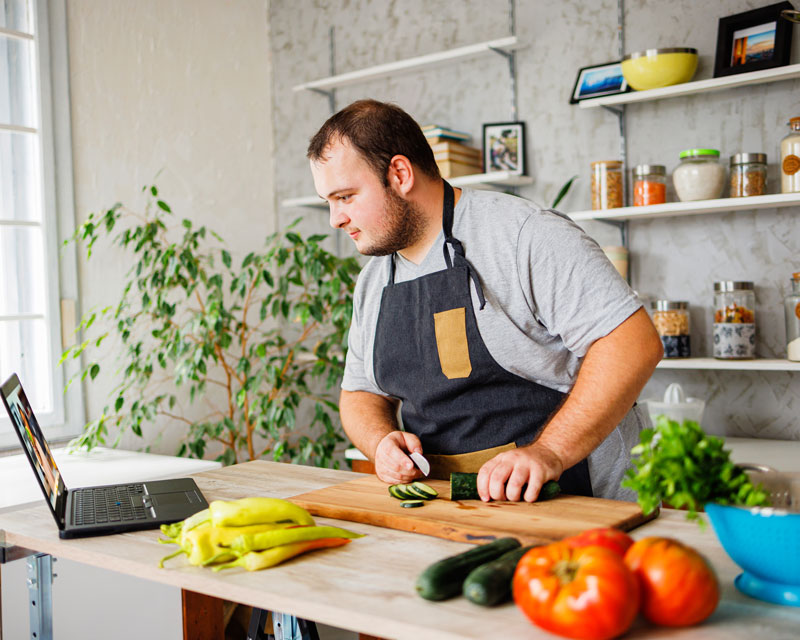 A man works in a kitchen preparing vegetables while following instructions on a laptop.