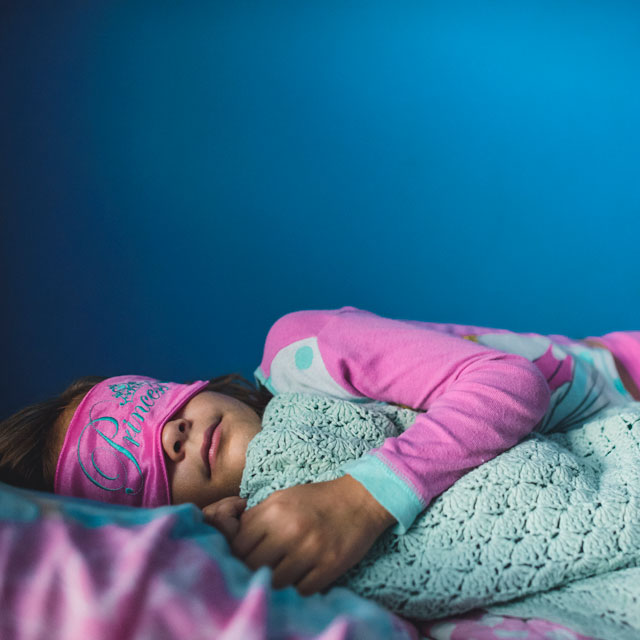 Grade-school-aged girl asleep holding a blanket and wearing a pink sleep mask.