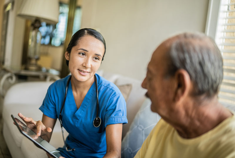Female nurse sits on a couch with an older male patient having a discussion while using a tablet.