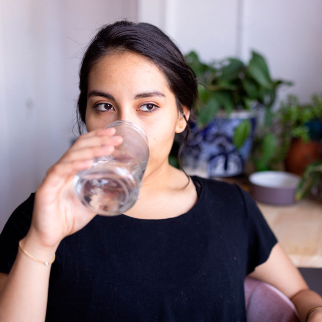 A Hispanic woman in a black T-shirt drinks water from a large glass.