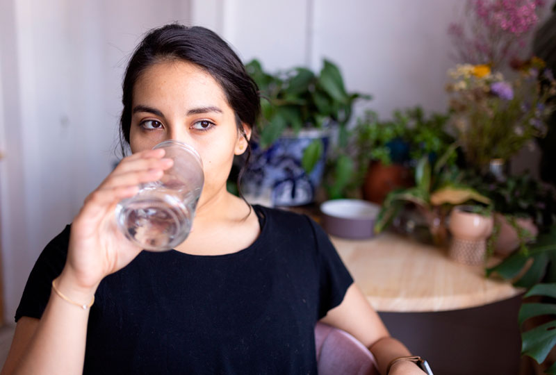 A Hispanic woman in a black T-shirt drinks water from a large glass.