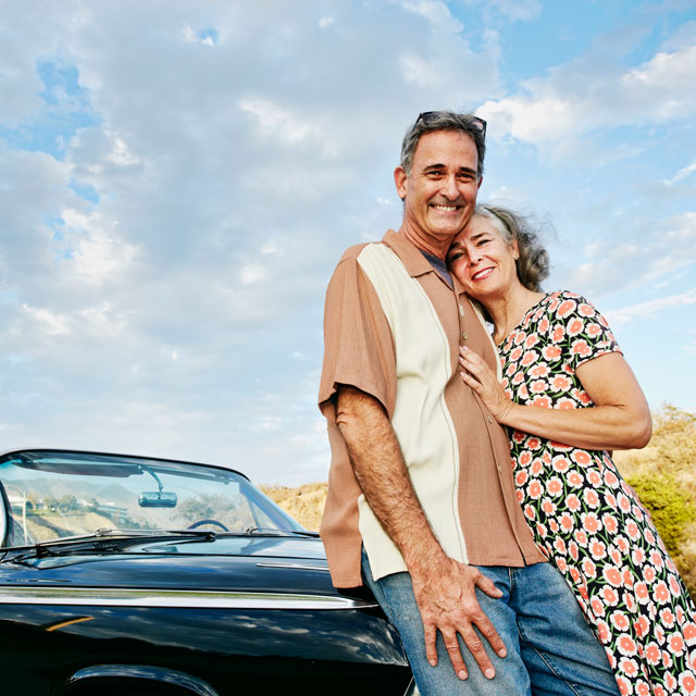 Older couple stand leaning against the hood of a vintage convertible car.