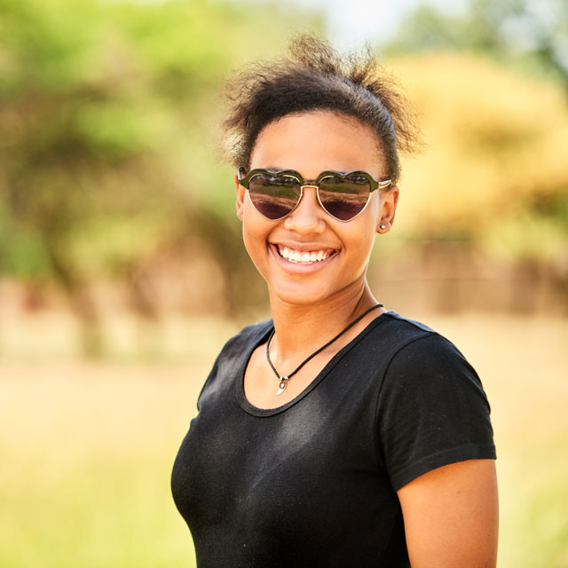 A young woman stands outdoors wearing a black T-shirt and heart-shaped sunglasses.