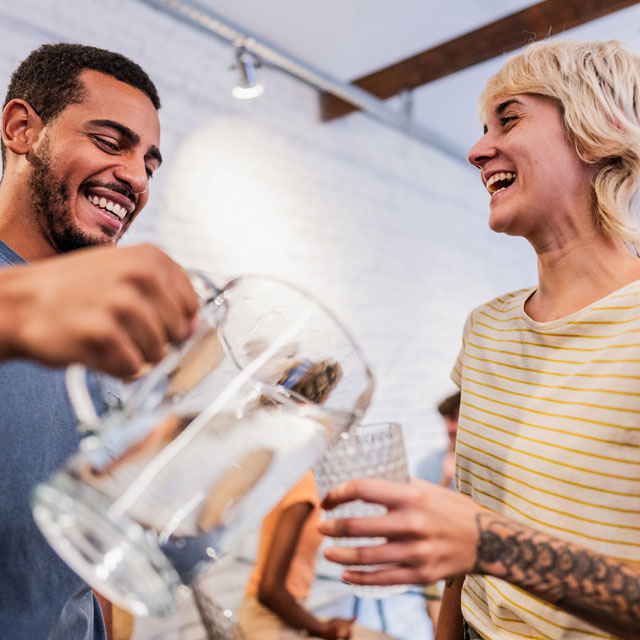 A young man pours water from a pitcher into a drinking glass for a. young woman.