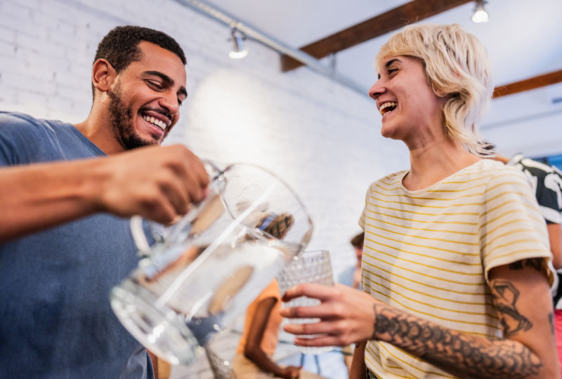 A young man pours water from a pitcher into a drinking glass for a. young woman.