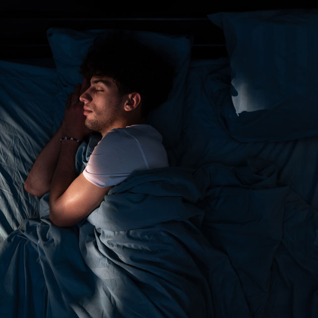 A young man sleeps in bed in a dark room curled up in a comforter.