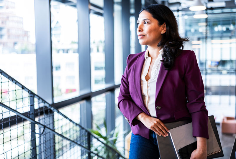 A woman who looks to be in her late 30s pauses on a stair landing to look out a window.