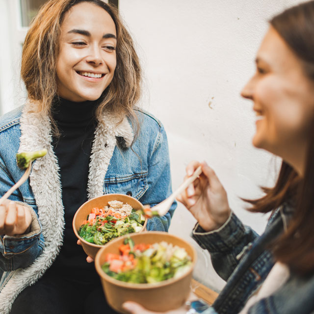 Two young women sitting together eating salad and veggie bowls.