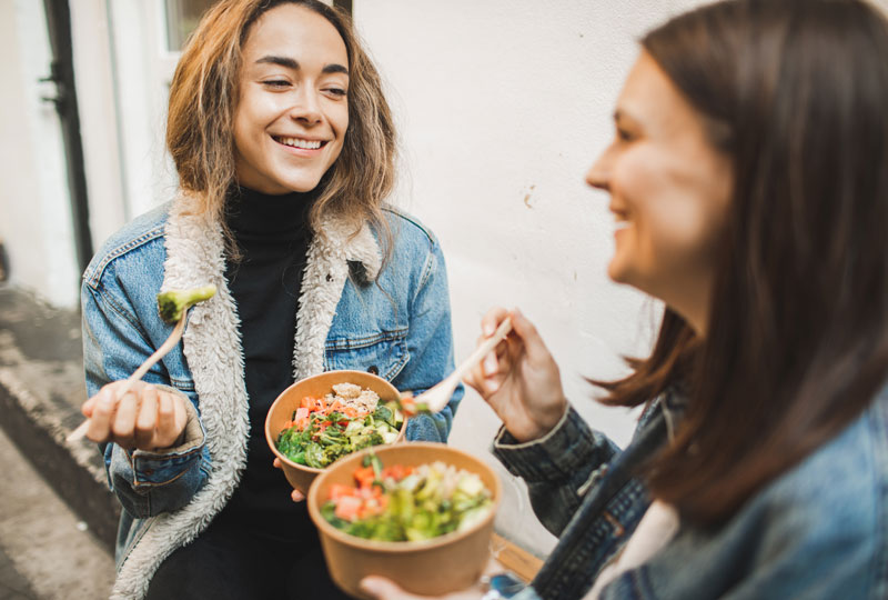 Two young women sitting together eating salad and veggie bowls.