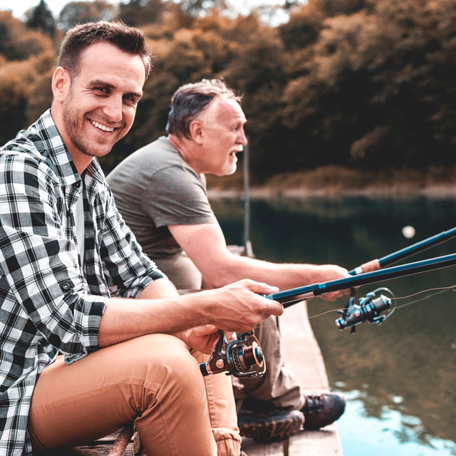 Two men, one middle aged and one older, sit on a dock, fishing in a river.
