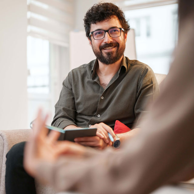 A man faces the camera and a counselor in a brightly lit office; the counselor is blurry in the foreground.