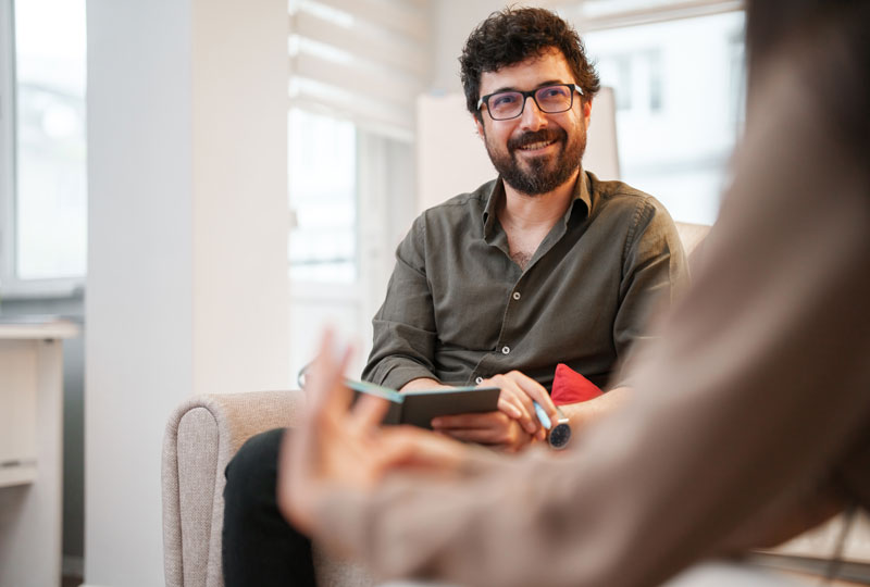 A man sits facing the camera and a counselor in a brightly lit office; the counselor is blurry in the foreground.